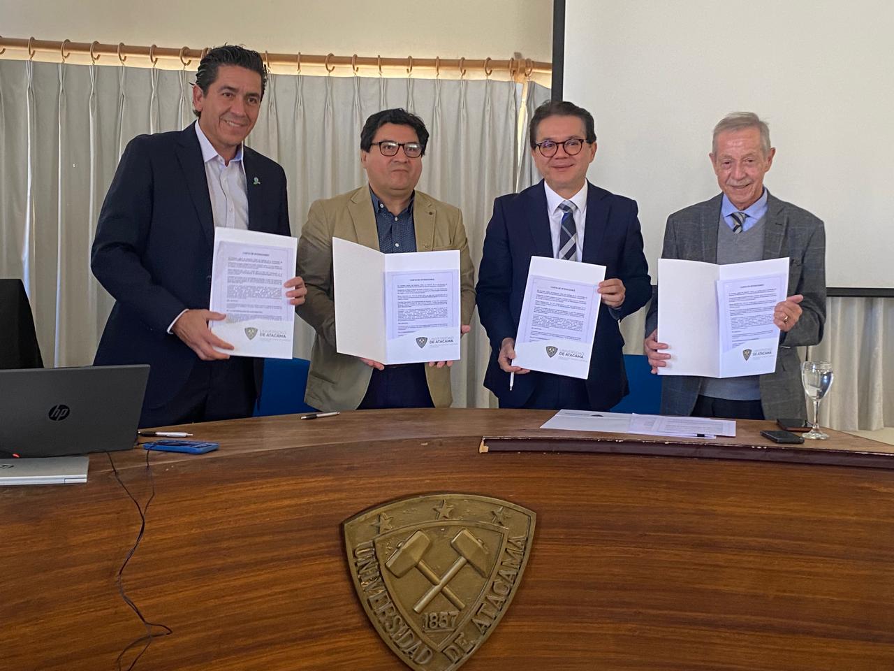 Cuatro personas sosteniendo firmas de convenio, de pie frente a una mesa de madera. Colaboración de compañía de aguas del Estado de Caera con la Universidad de Atacama