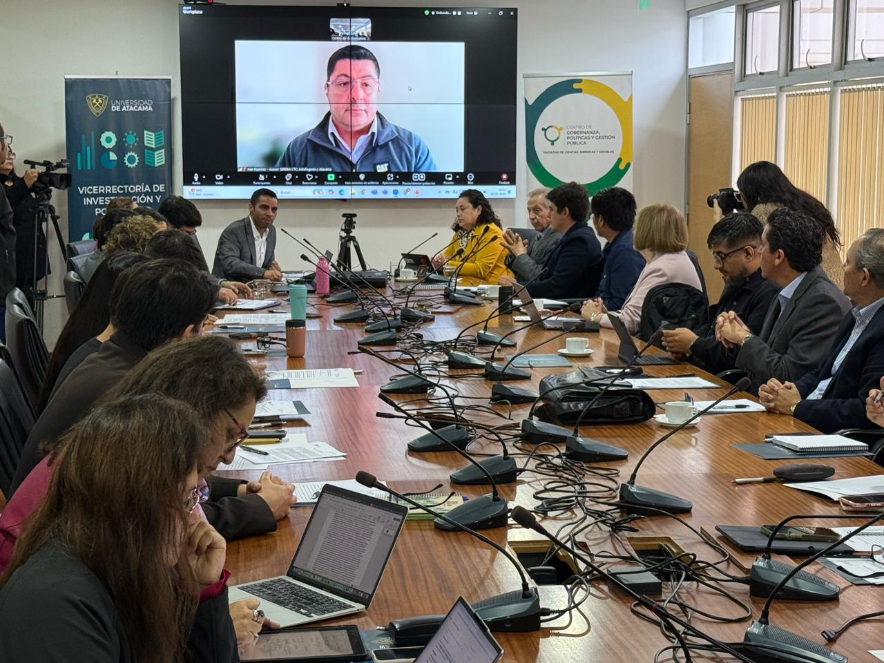 Sala de reuniones con muchos micrófonos en su superficie, con personas sentadas al rededor. En la cabecera, la imagen proyectada de un hombre que asiste telemáticamente. A sus costados, dos pendones institucionales. Luz blanca y mesa de madera.