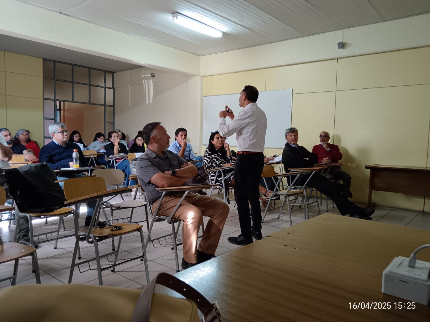 Una sala de clases de paredes crema con un foco de luz fría en el techo. Un profesor con camisa blanca dando una cátedra a otros profesores, sentados en sillas universitarias.